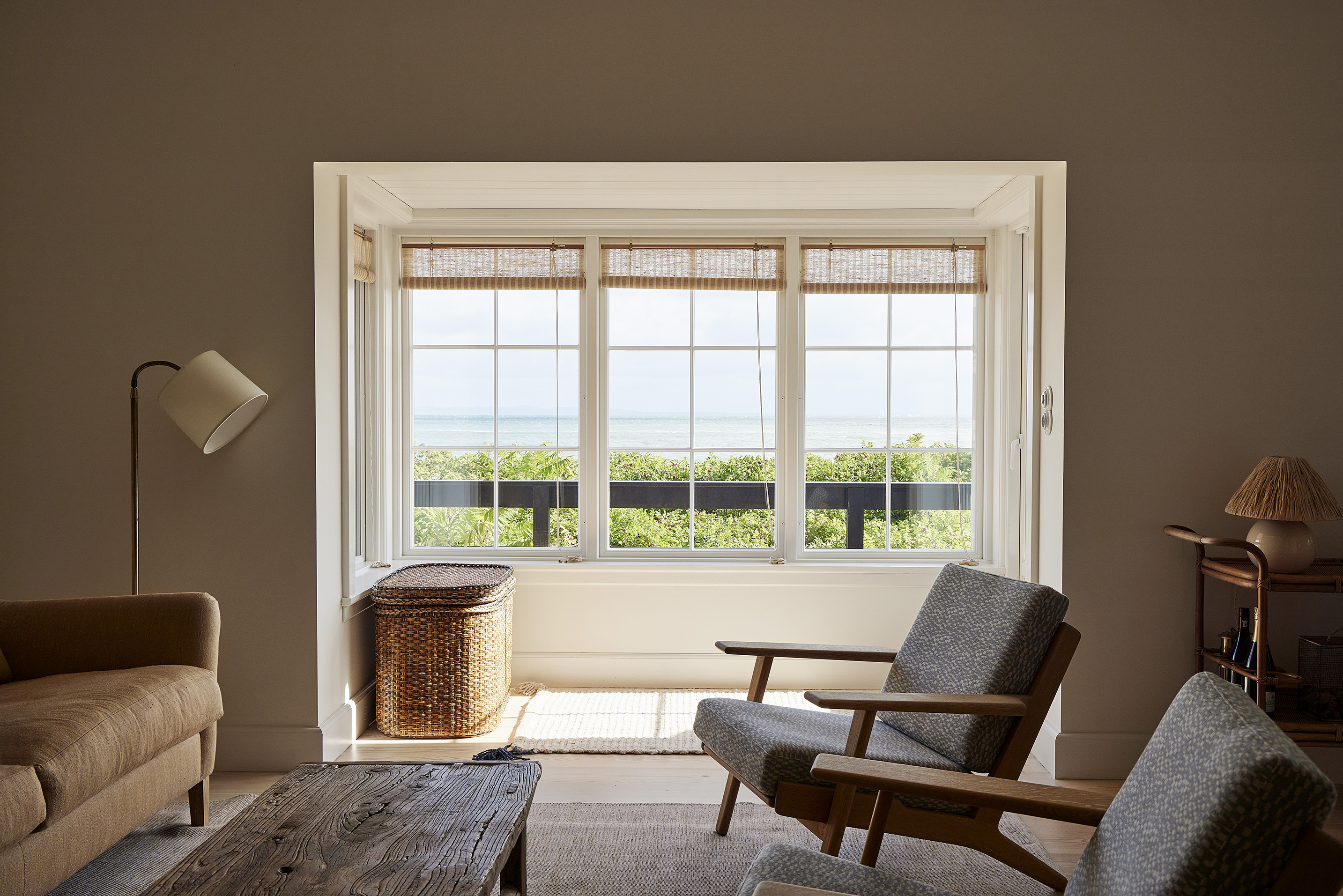 scandinavian coastal style living room with two gray and timber armchairs, timber coffee table, brown leather sofa, rattan basket, floor lamp, and framed windows