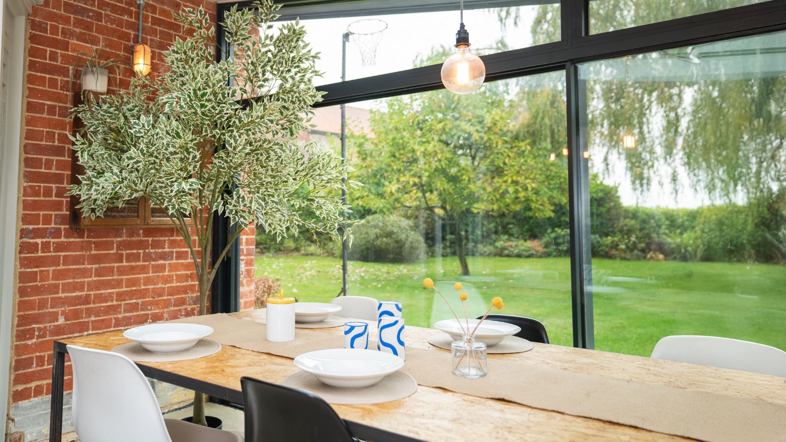 Dining area with large windows revealing outdoor lawn and trees. Pictured inside is a large indoor tree, a brick wall, a wooden dining table with bowls and placements, and black-and-white chairs.