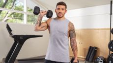 Man wearing gray vest holds dumbbell by his shoulder. A treadmill and other gym equipment is behind him.