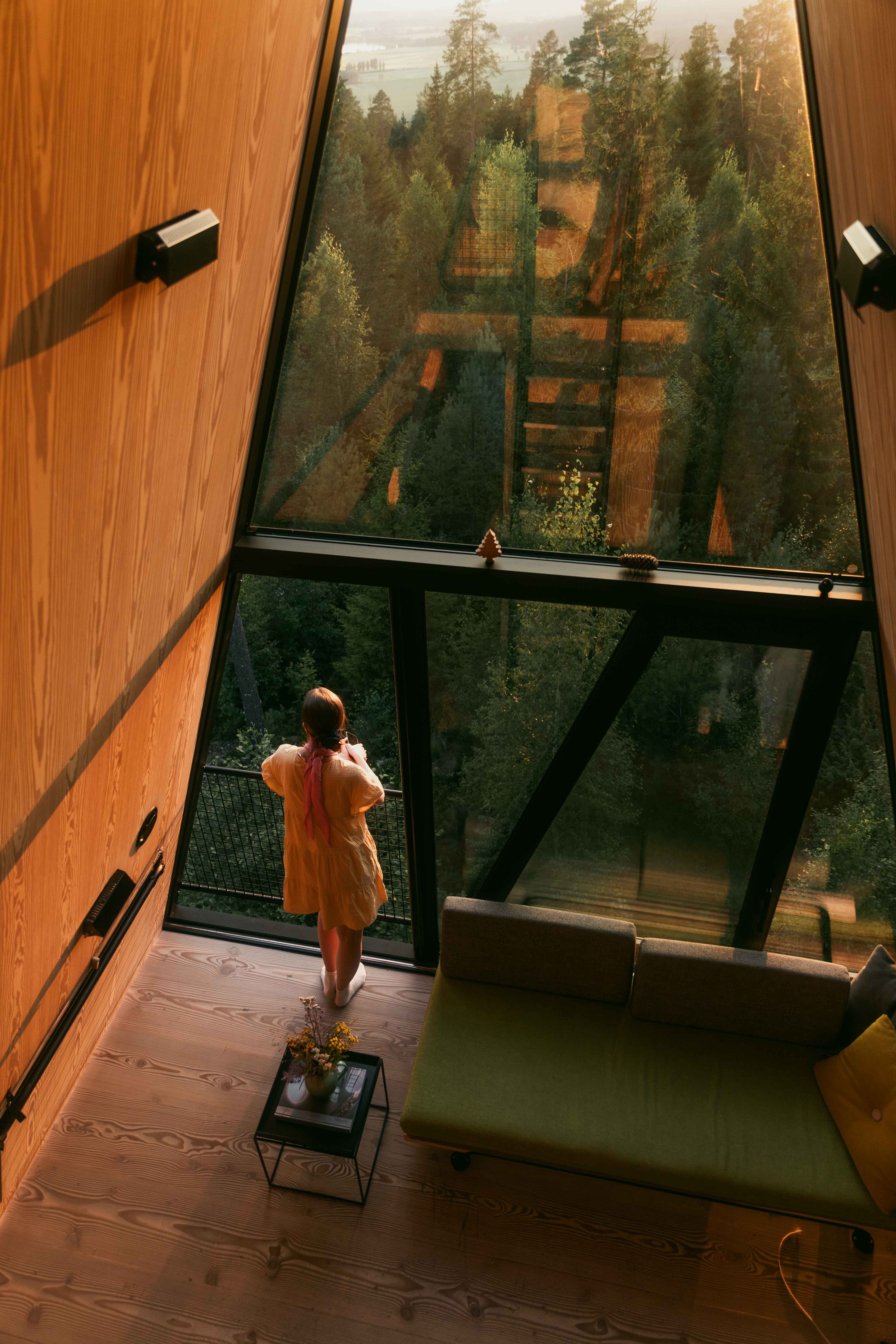 The interiors of a design cabin in the woods are captured at sunset as one of its guests stares outside of its triangular glassy facade.