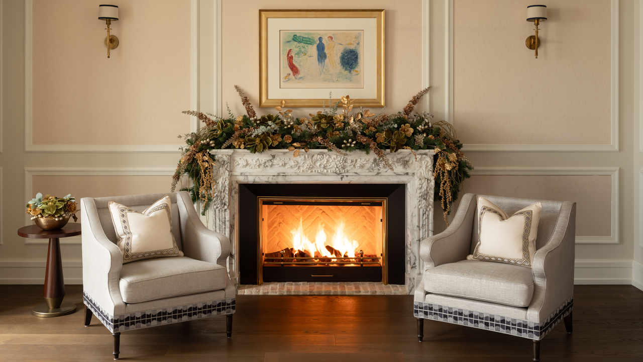 A living room area with a fireplace in the middle of two arm chairs and a garland above the fireplace as well as framed artwork