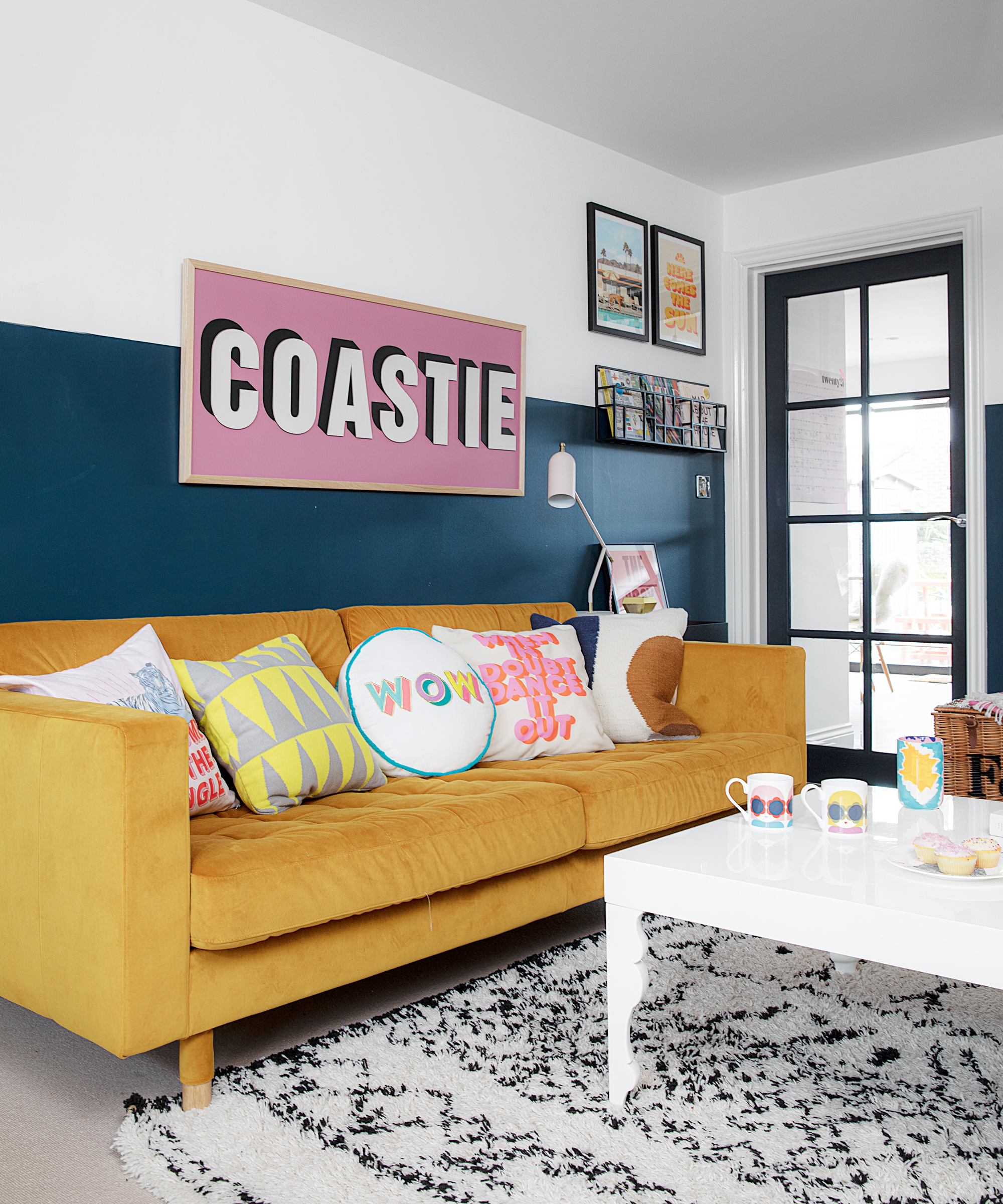 Yellow sofa with assorted, playful scatter pillows, two tone painted wall, tactile patterned mono rug, and large typo sign on wall.