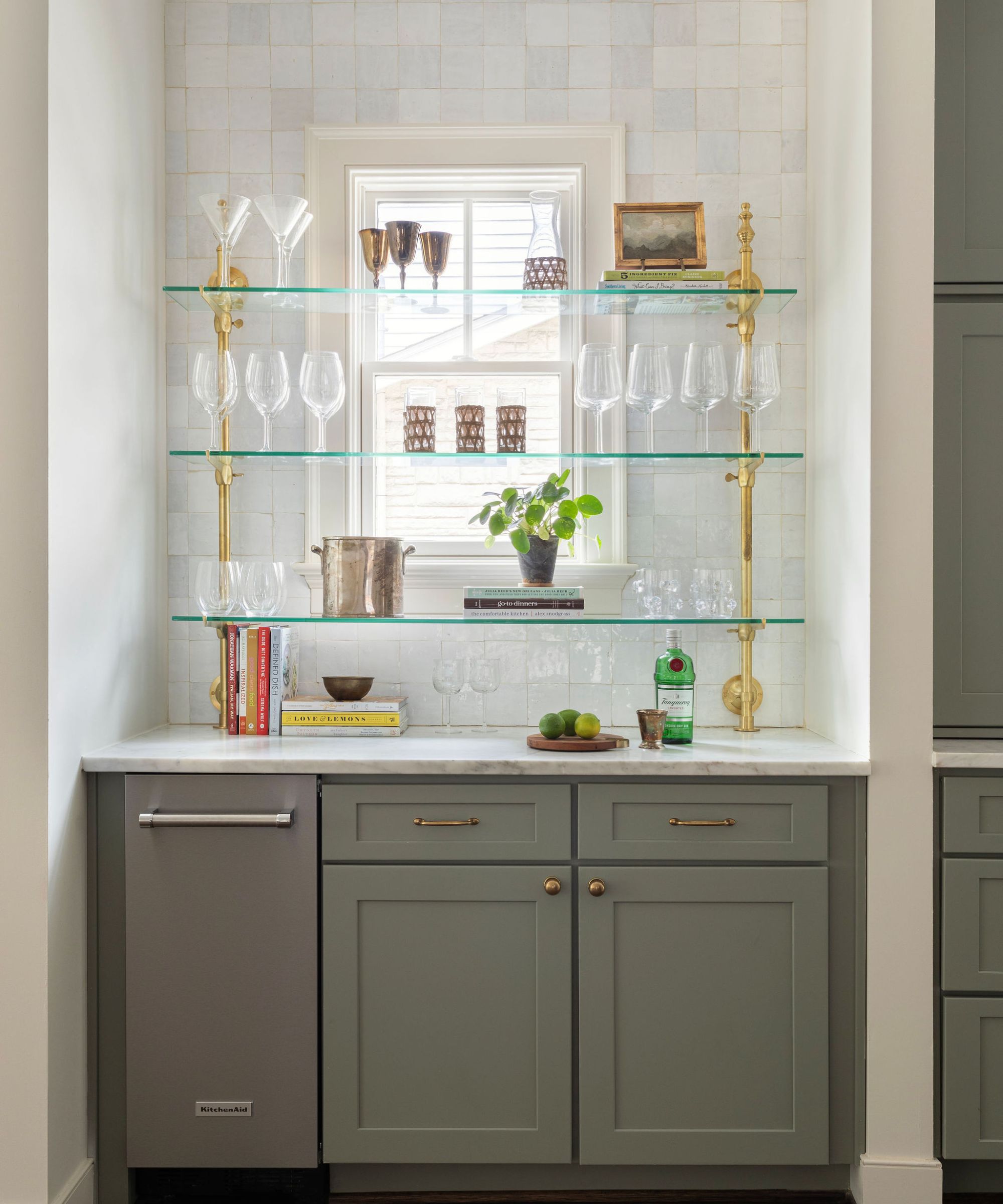 Home bar with white tiles, grey painted cabinetry, glass shelves with gold hardware, and a collection of wine glasses and barware