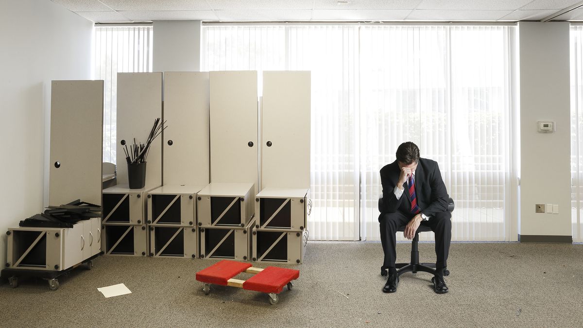 A businessman sitting on an office chair with his head in his hands in an office that&#039;s being emptied out because they have gone out of business