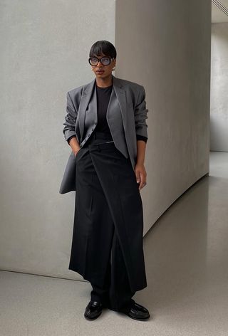 A woman wearing a gray blazer over gray waistcoat and black t-shirt styled with a black skirt layered over black trousers and black loafers.