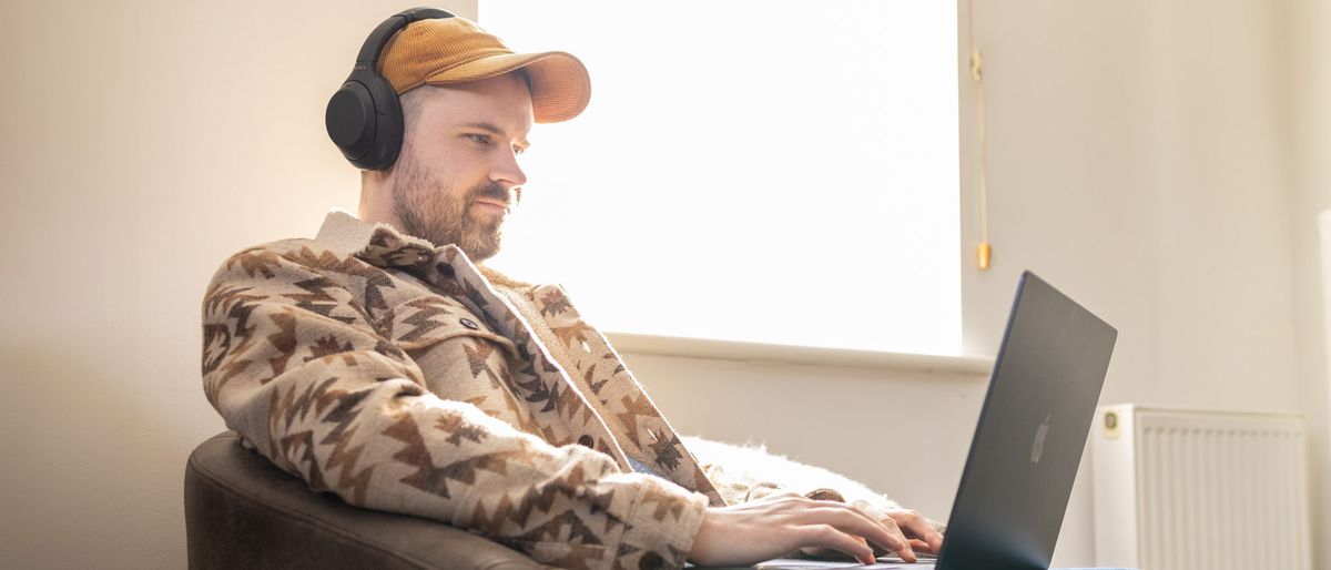 A man wearing a black pair of Sony WH-1000M4 headphones at home on a chair interacting with a MacBook on his lap