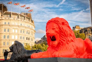 Alternative view of Please Feed The Lions by Es Devlin at Trafalgar Square during the day - a red lion statue on a platform with another bronze lion statue, a person and buildings in the background
