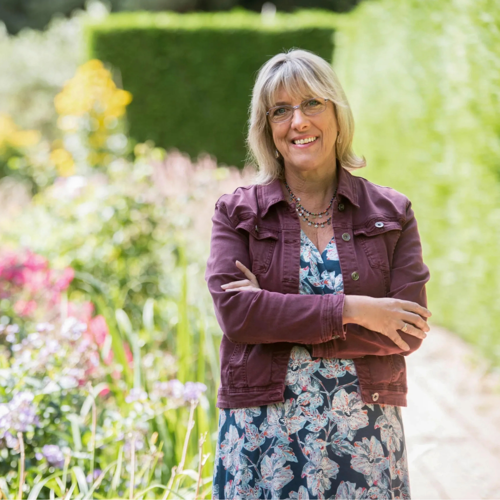 female with bobbed blond hair, wearing dark pink jacket and floral dress stood in garden