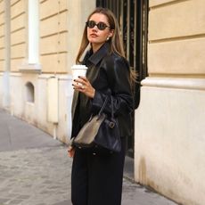 style influencer Lovia Barkman poses in Paris holding a coffee while wearing black oval sunglasses, a black leather jacket, black The Row bag, and black pants