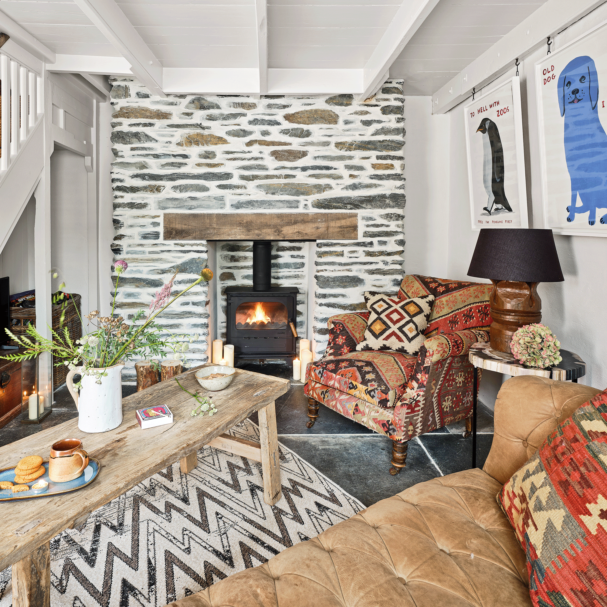 a living room with a large leather chesterfield style sofa, a rustic coffee table patterned armchair, rug and cushion and a stone fireplace with a wood burning stove