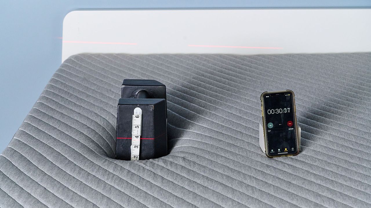 A close up of a 25kg weight balanced on the Siena Signature Mattress in our sleep studio. There's a tape measure attached to the side of the weight and a laser line is hitting the tape measure at roughly 3.5 inches, indicating how far the weight has sank into the mattress. To the right, a phone displaying a stopwatch at 30 seconds; how long the weight has been on the Siena Mattress.