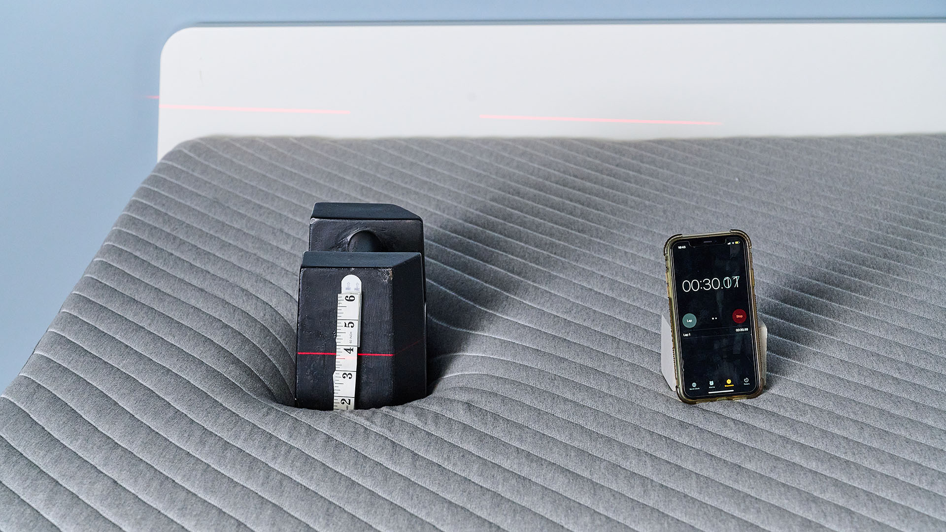 A close up of a 25kg weight balanced on the Siena Memory Foam Mattress in our sleep studio. There&#039;s a tape measure attached to the side of the weight and a laser line is hitting the tape measure at roughly 3.5 inches, indicating how far the weight has sank into the mattress. To the right, a phone displaying a stopwatch at 30 seconds; how long the weight has been on the Siena Mattress.