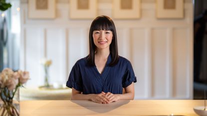 Marie Kondo sat at a wooden table with a white wall behind her, in her Netflix show Sparking Joy, to illustrate Marie Kondo tips for organizing a closet