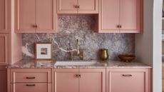 A close-up image of kitchen cabinetry. The cabinets are a dusty pink color and there is gray marble on the countertops as well as the back splash