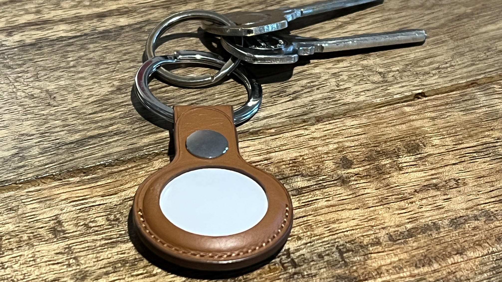 an Apple AirTag on a brown leather keyring, attached to some keys on a wooden surface