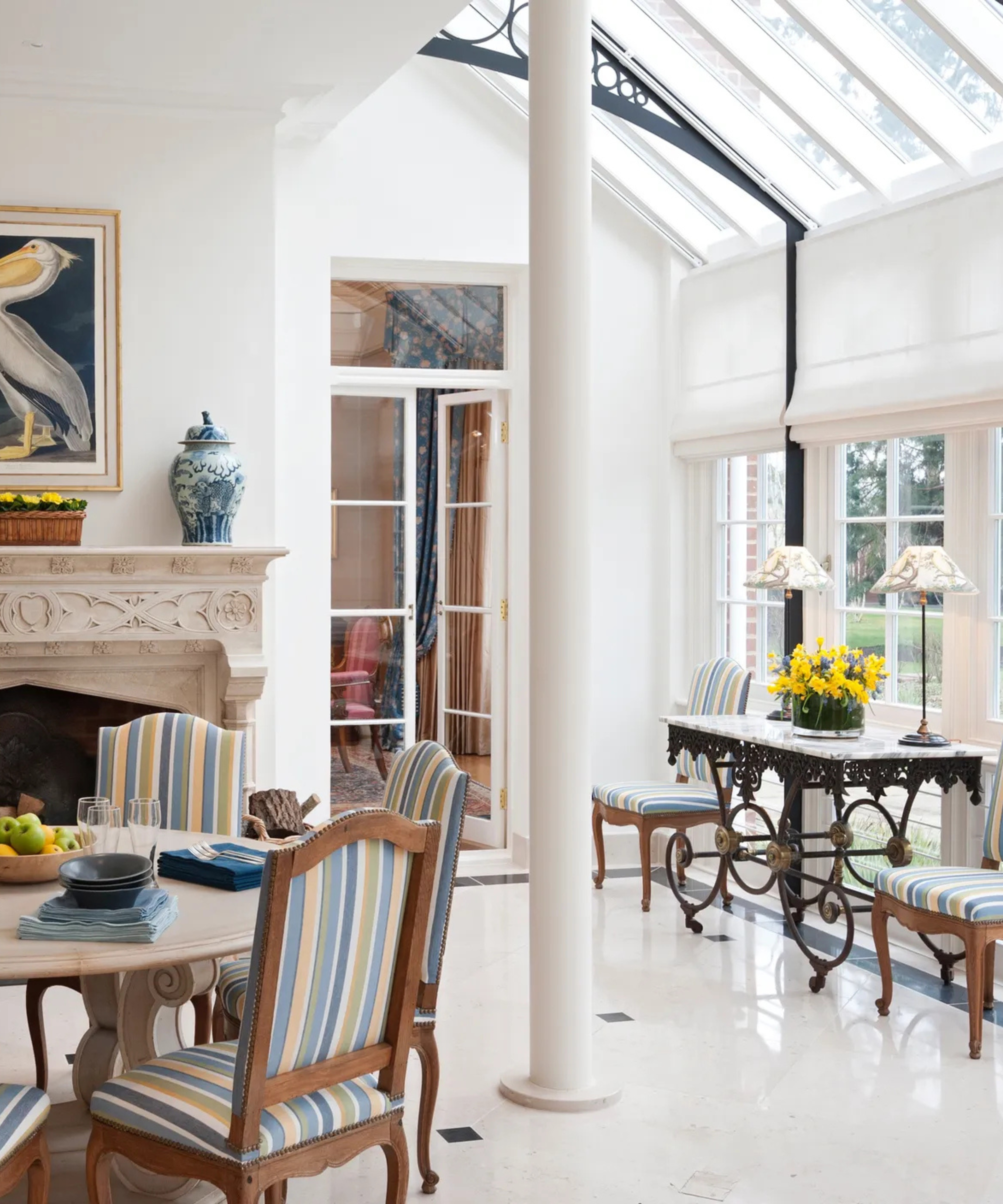 A bright conservatory with automated Roman roof blinds, a glazed ceiling, striped dining chairs, a carved stone fireplace and large windows overlooking the garden.