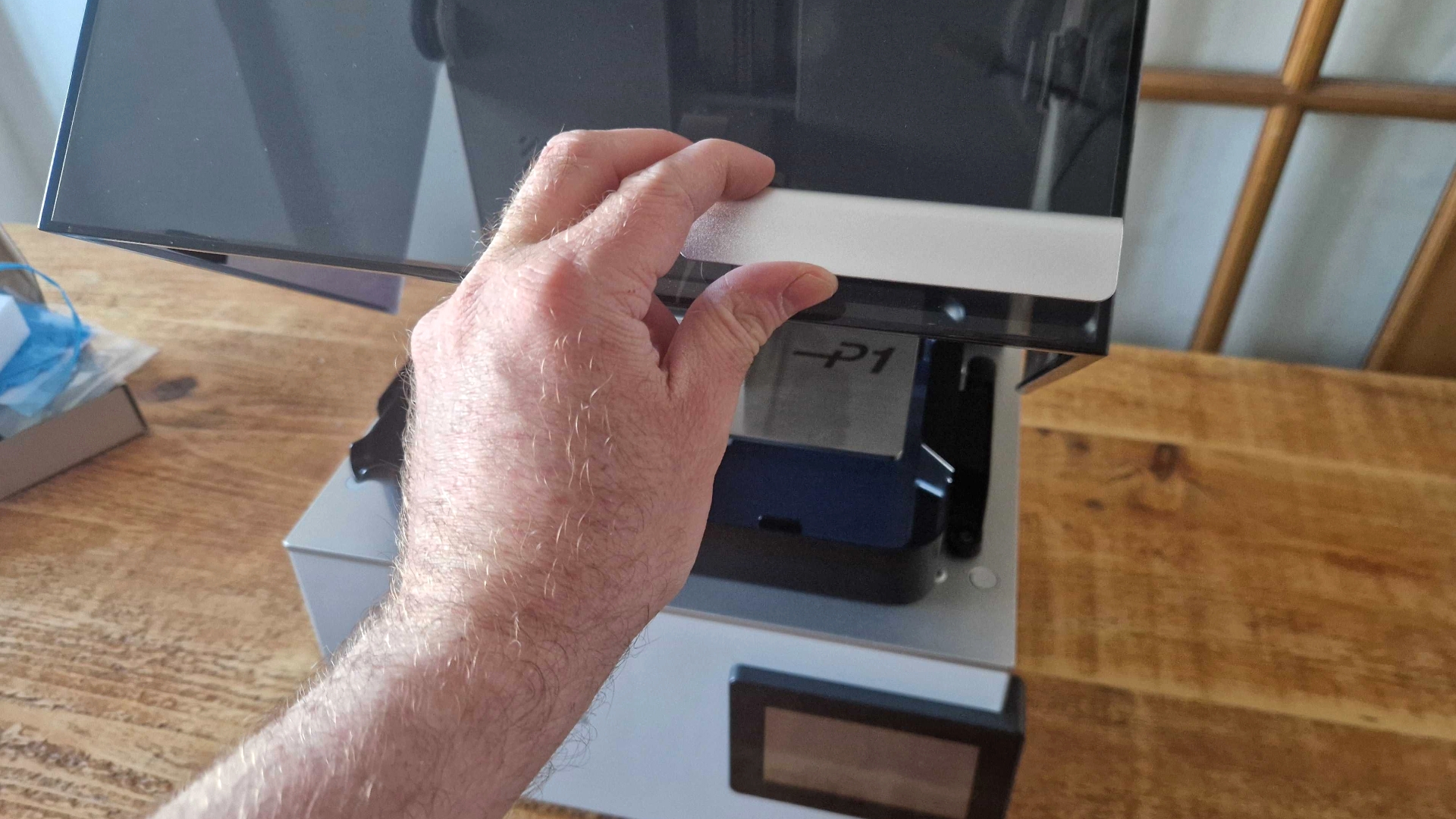 A hand lifts up the lid for the Anycubic Photon P1, which sits on a wooden table