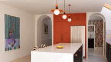 A bright kitchen with an orange accent storage unit by a silver fridge, a stone island with a bowl of lemons, a pair of zebra print seats, three pendant orange lights, a framed painting and arched doorways leading to a room with floral wallpaper