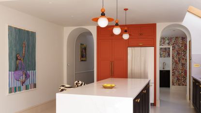 A bright kitchen with an orange accent storage unit by a silver fridge, a stone island with a bowl of lemons, a pair of zebra print seats, three pendant orange lights, a framed painting and arched doorways leading to a room with floral wallpaper