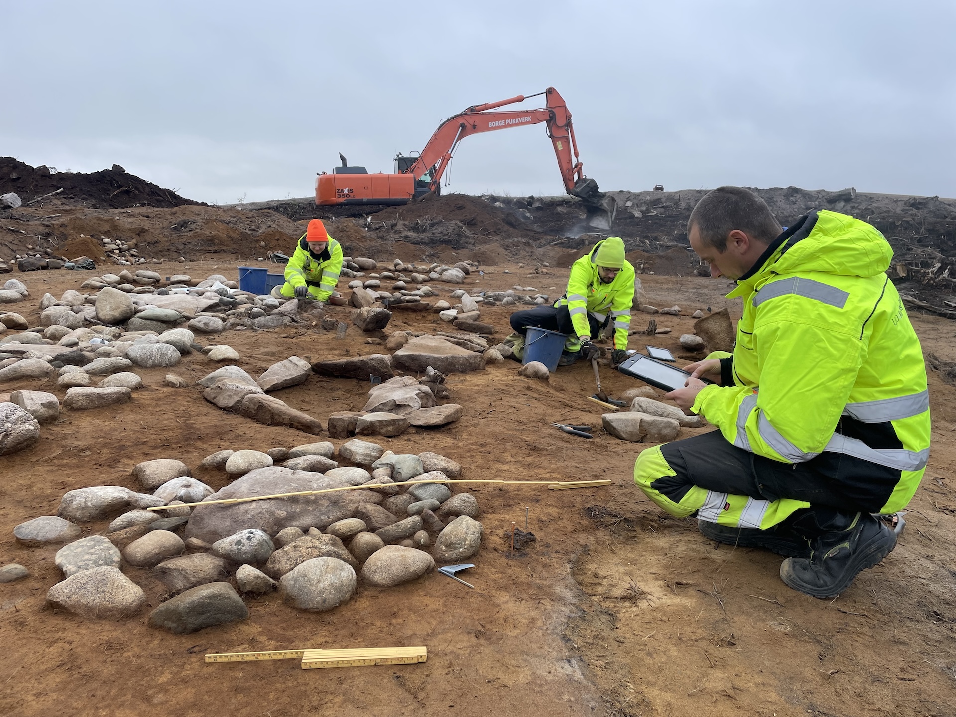 A photo showing archaeologists working on the graves with a bulldozer in the background