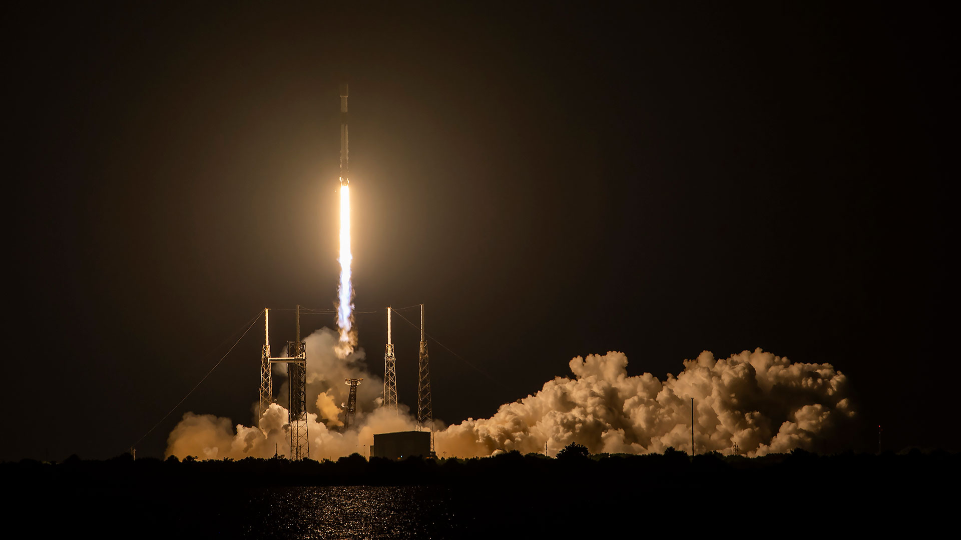 a white and black rocket rides a pillar of orange, blue and white flame off its launch pad into the night sky.