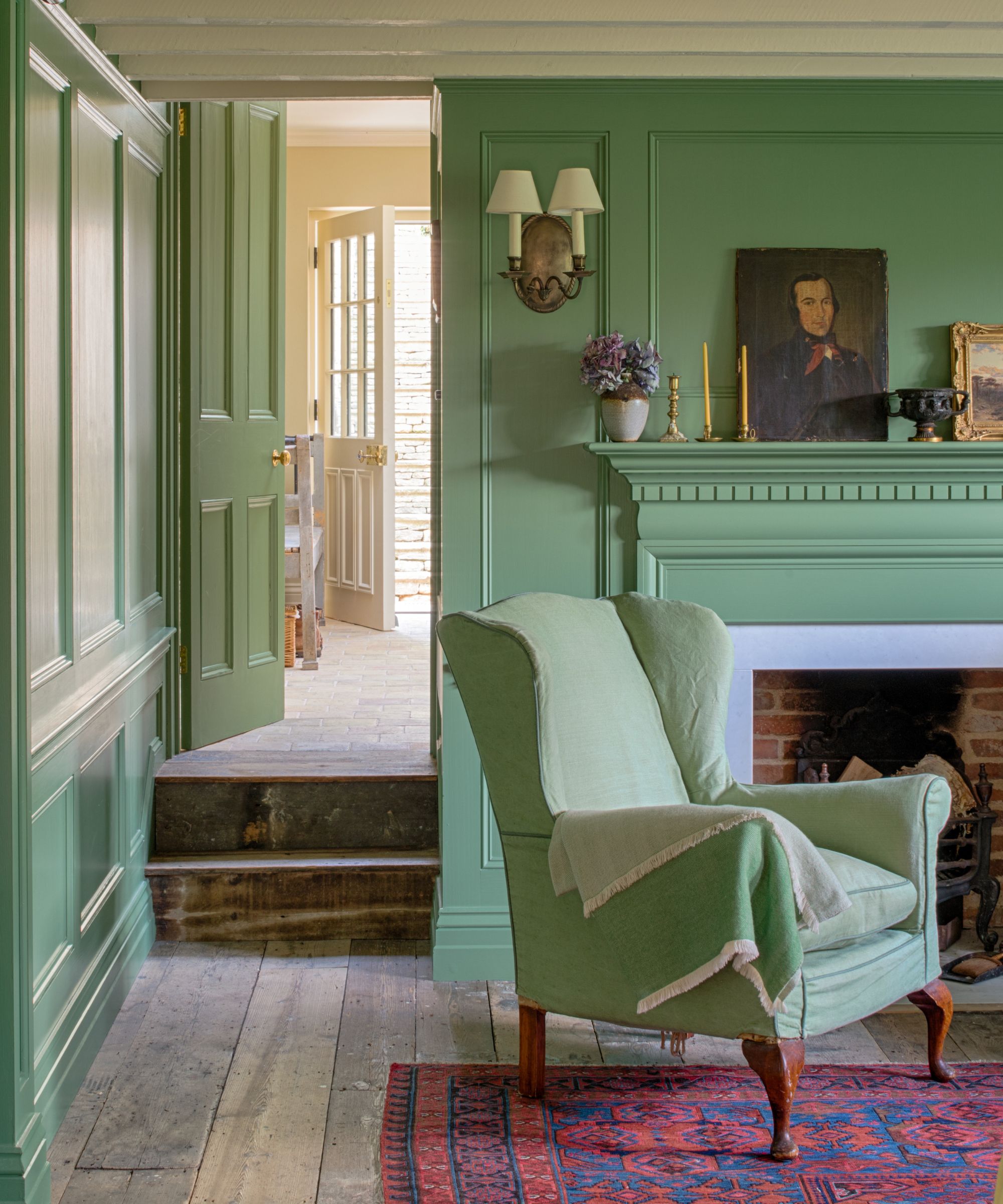 Dining room with Calke green by Farrow &amp;amp; Ball painted walls, and door, wooden floorboards, a red Persian rug, a pale green armchair, and a fireplace decorated with antique art and candlesticks