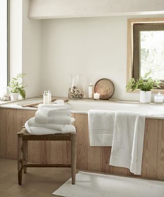 Guest bathroom with white and wood, built in bath with wooden panels, stool with white towels, plants, toiletries