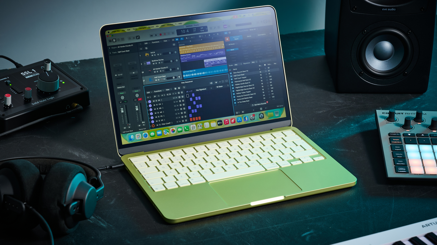 An Apple MacBook Neo on a desk with an audio interface, studio monitors, headphones, and two MIDI controllers. Displayed on the screen is the Logic Pro DAW