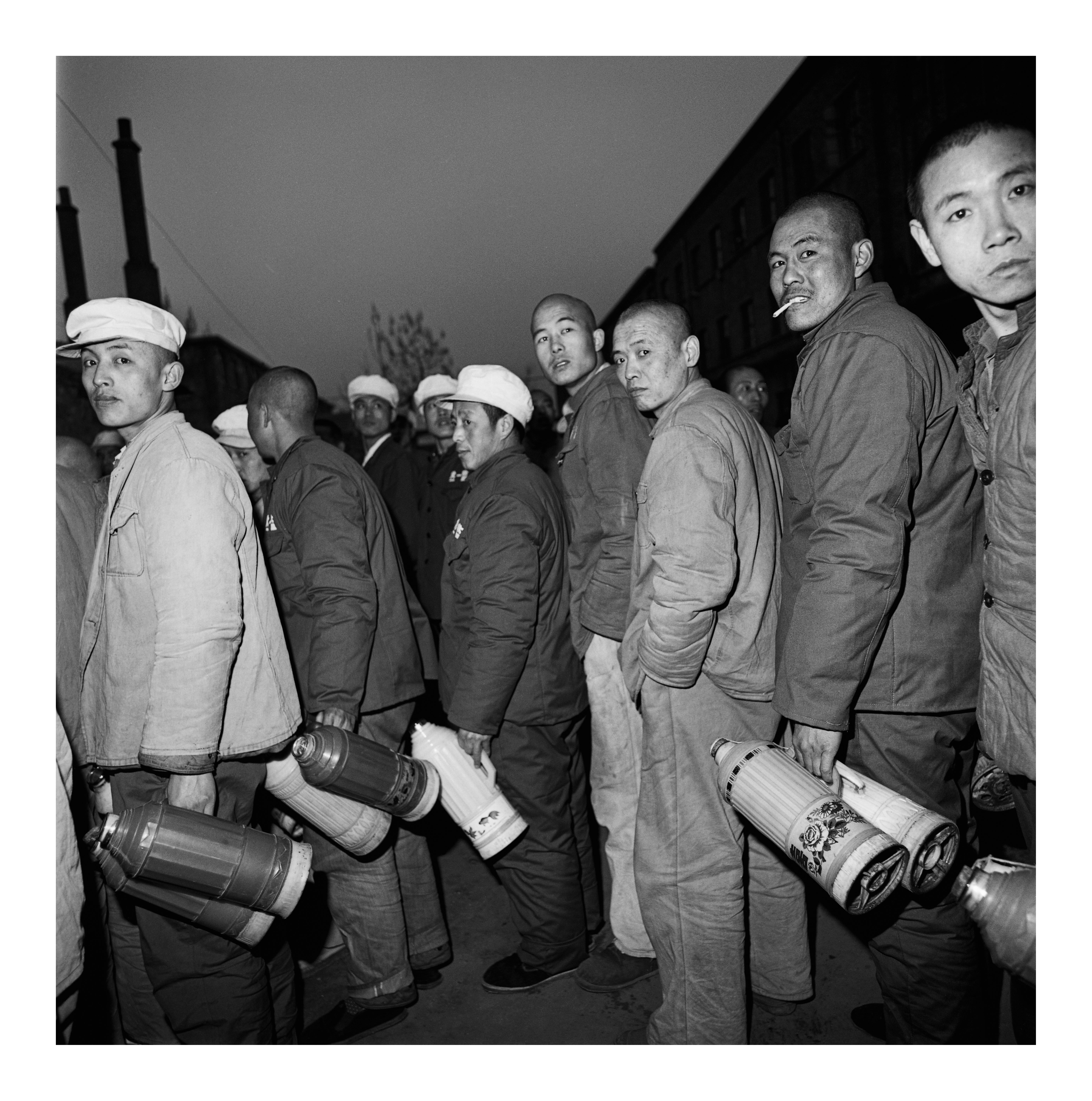 A black-and-white photograph capturing a tight line of men in work clothes and caps looking back toward the camera, with several of them holding large, insulated thermoses.