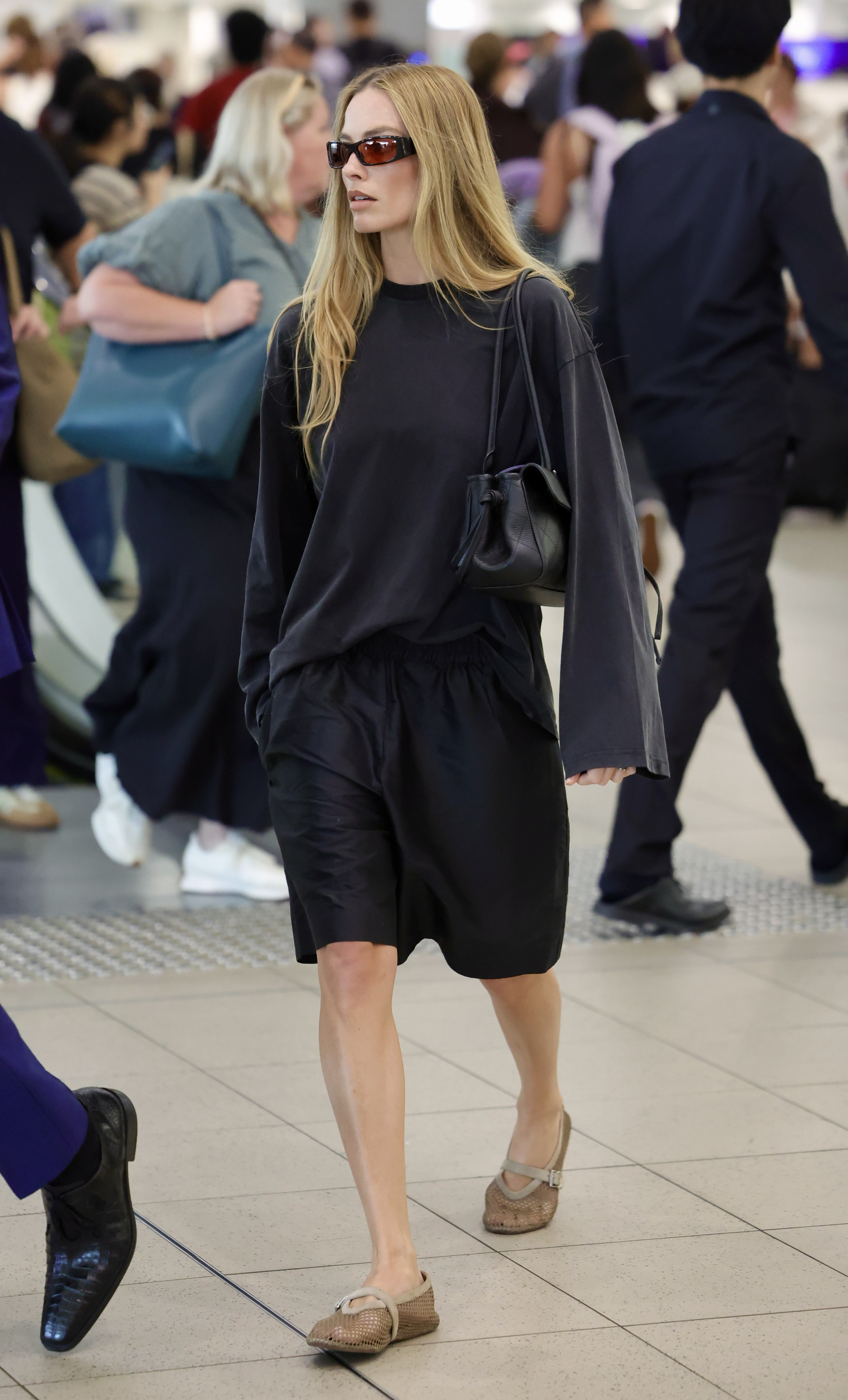 An image of Margot Robbie at the airport wearing a black long-sleeve shirt, tailored trousers and mesh flats.