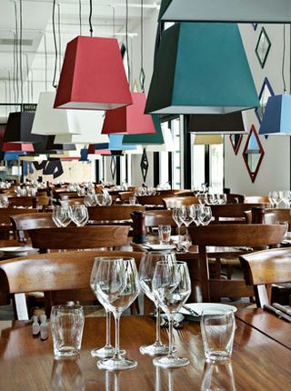 Inside a restaurant, close up to the wood tables and glassware ontop and multi-coloured lamp shades hanging from above.