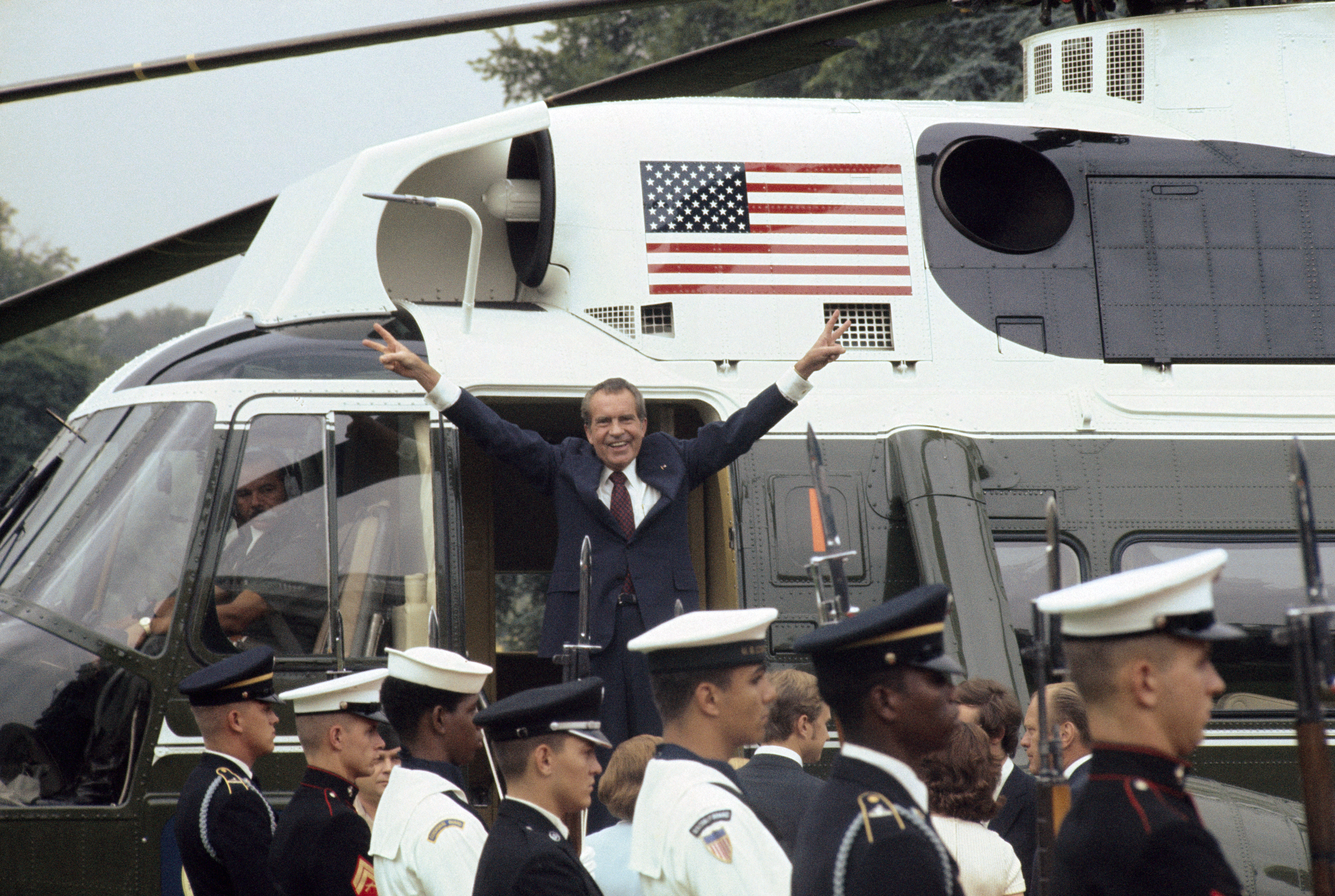 Richard Nixon throwing his arms in the air getting out of a helicopter with soldiers below him