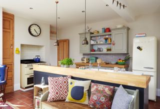Open-plan freestanding kitchen, with an aga to the left, a white smeg fridge to the right, a built in gray and wood unit at the back and a freestanding island, behind a seating area with lots of decorative colorful pillows