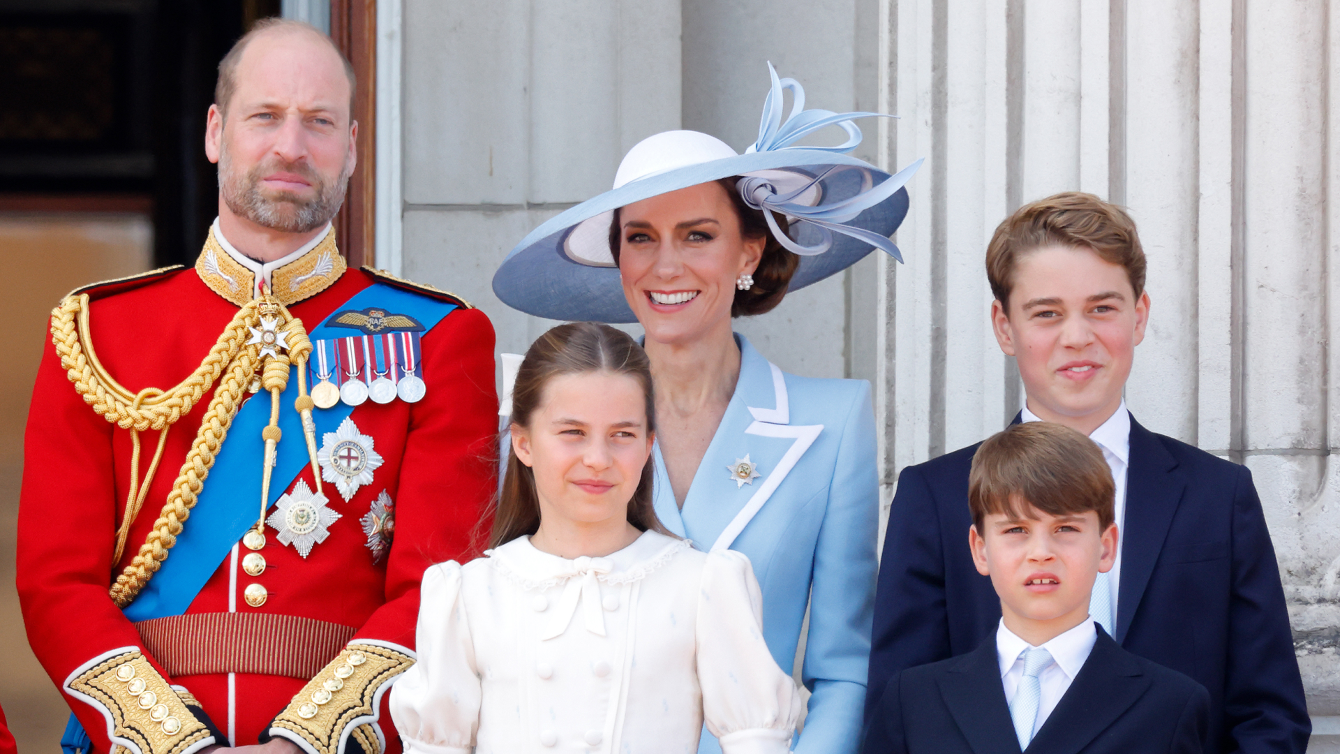 Prince William wears red military uniform and has a bald head and a beard, and stands next to wife Princess Middleton who wears a baby blue coat dress and matching hat, and their three kids