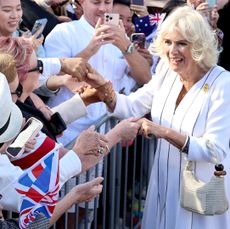 Queen Camilla wearing a white dress and shaking hands with a large crowed of fans waving British and Australian flags