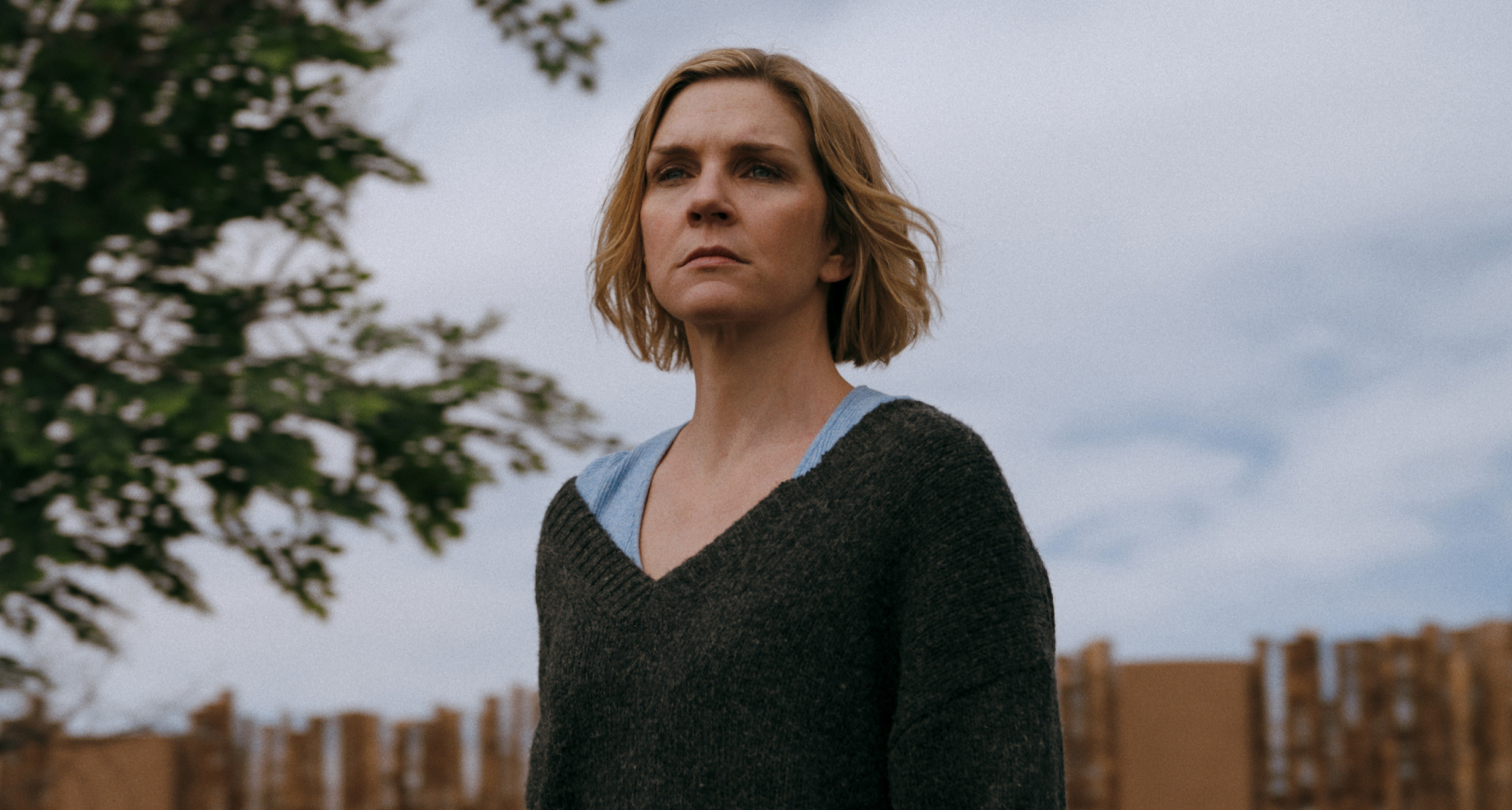 rhea seehorn as carol wearing a grey sweater standing in her yard and staring into the distance in pluribus season 1