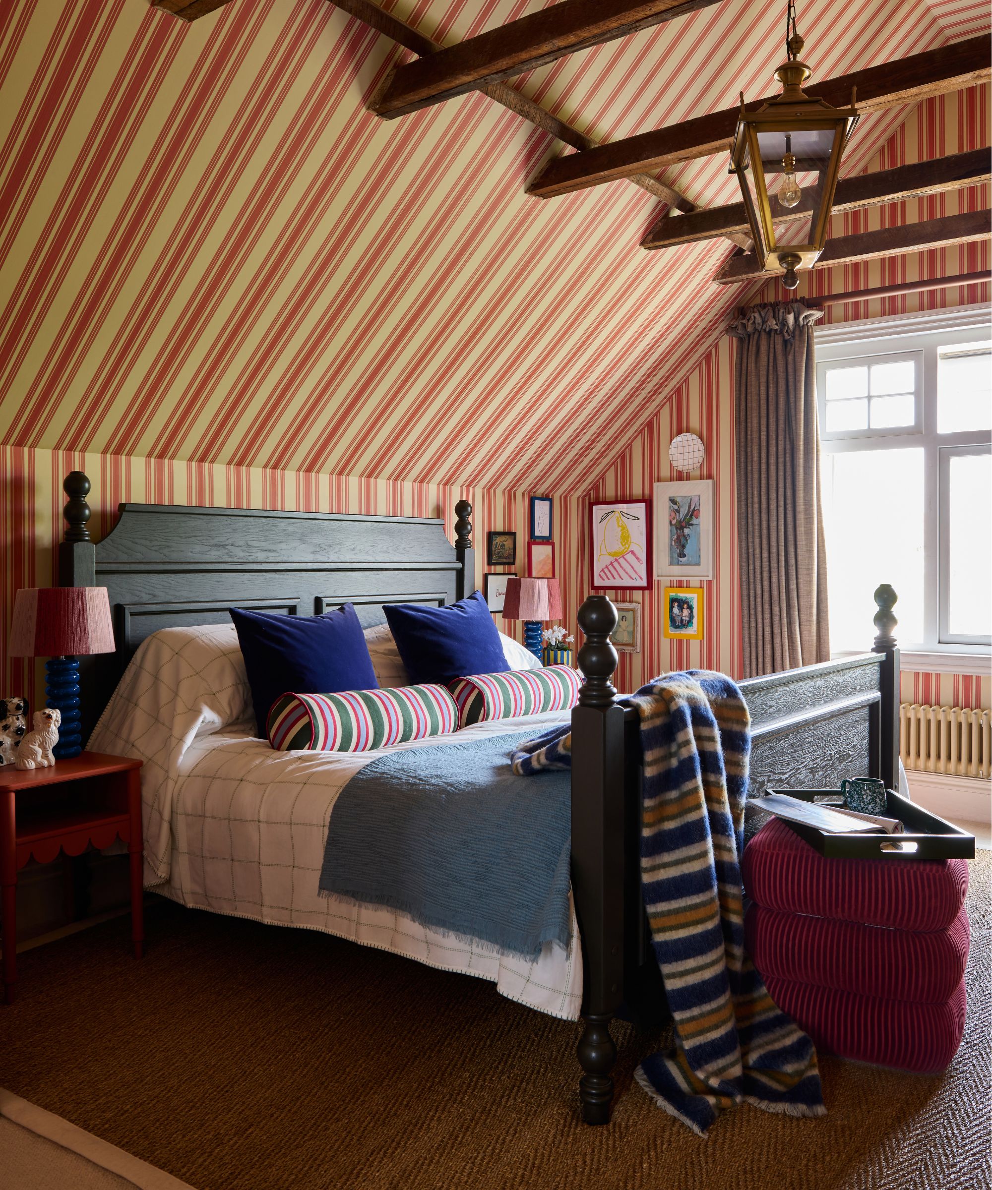 bedroom with a sloped ceiling and beige and red striped wallpaper that goes up and over the ceiling. A large dark wooden vintage bed sits in the middle styled with striped throws and pillows with a velvet stool at the foot of the bed