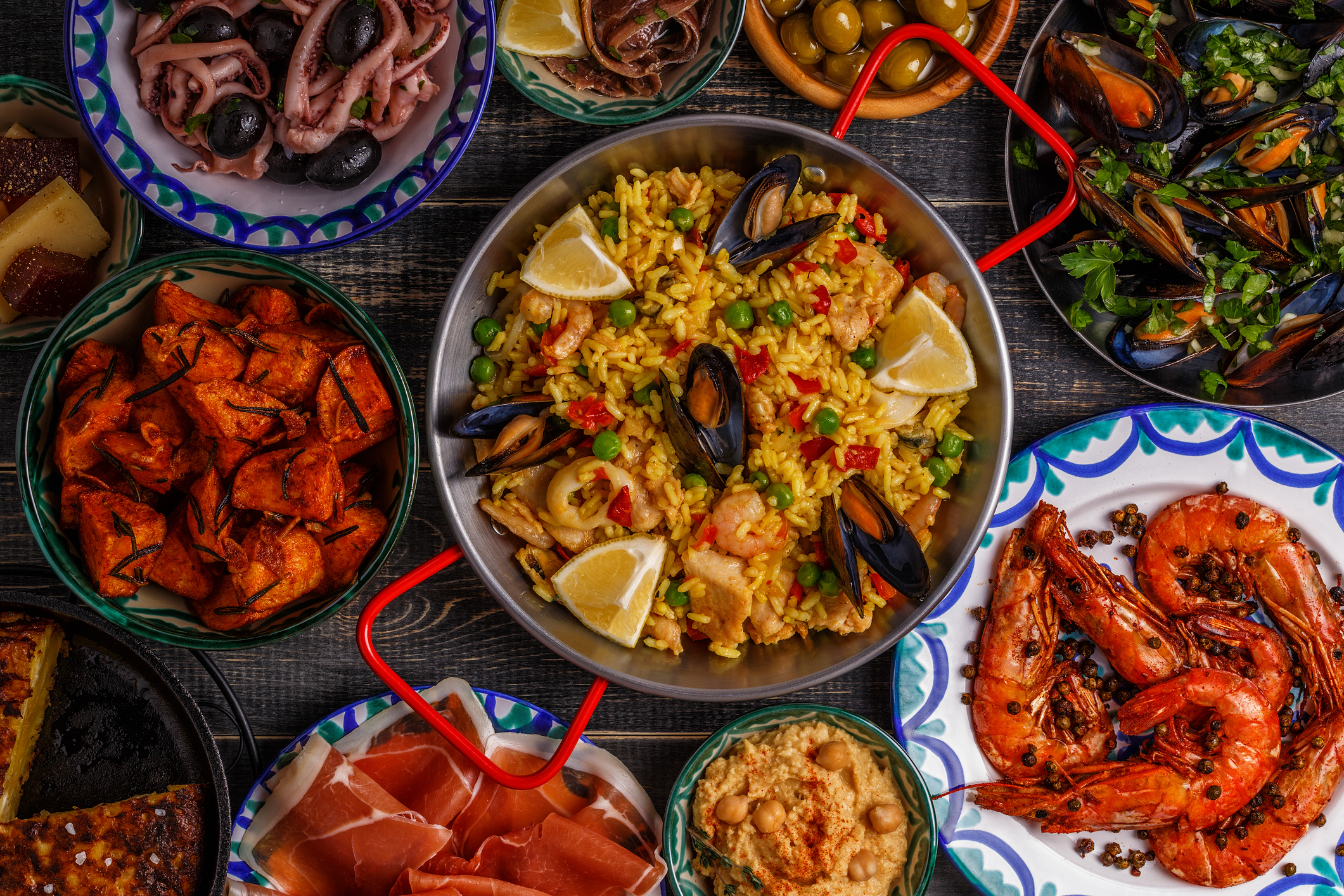 Typical spanish tapas, including slices jamon, bowls with olives, anchovies, spicy potatoes, mashed chickpeas, shrimp, calamari, manchego with quince marmalade (membrillo), pans with tortilla, paella, mussels on a wooden table.