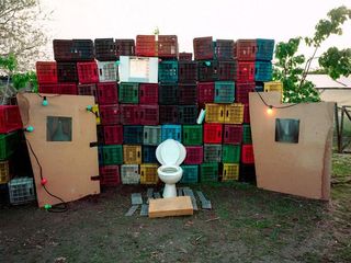 Outside stage setting. A toilet with its seat lifted sits in the centre of the stage with empty crates piled up high to create a colourful backdrop.