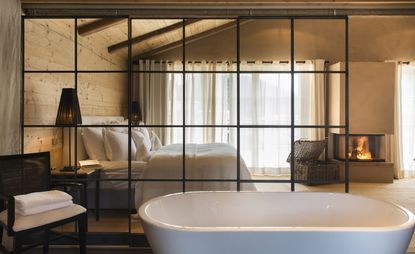 Interior view of a room at San Luis featuring a large window with white curtains, fireplace, bed with white pillows and linen and an area with a white bath separated by a black grid style partition