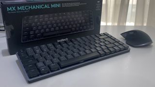 Det trådlösa tangentbordet Logitech MX Mechanical Mini står på ett vitt skrivbord bredvid sin tillhörande förpackning.
