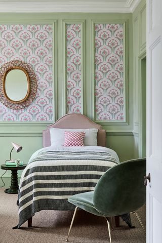 pink and green guest bedroom with pink and green stylised floral wallpaper within apple green painted paneling, dark green modern side table, velvet armchair, wicker mirror, pink upholstered bed