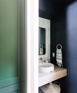 A small bathroom with a dark green wall and a dark blue wall, and a white rectangular mirror and a white sink on a light wooden shelf with a gray storage basket below it