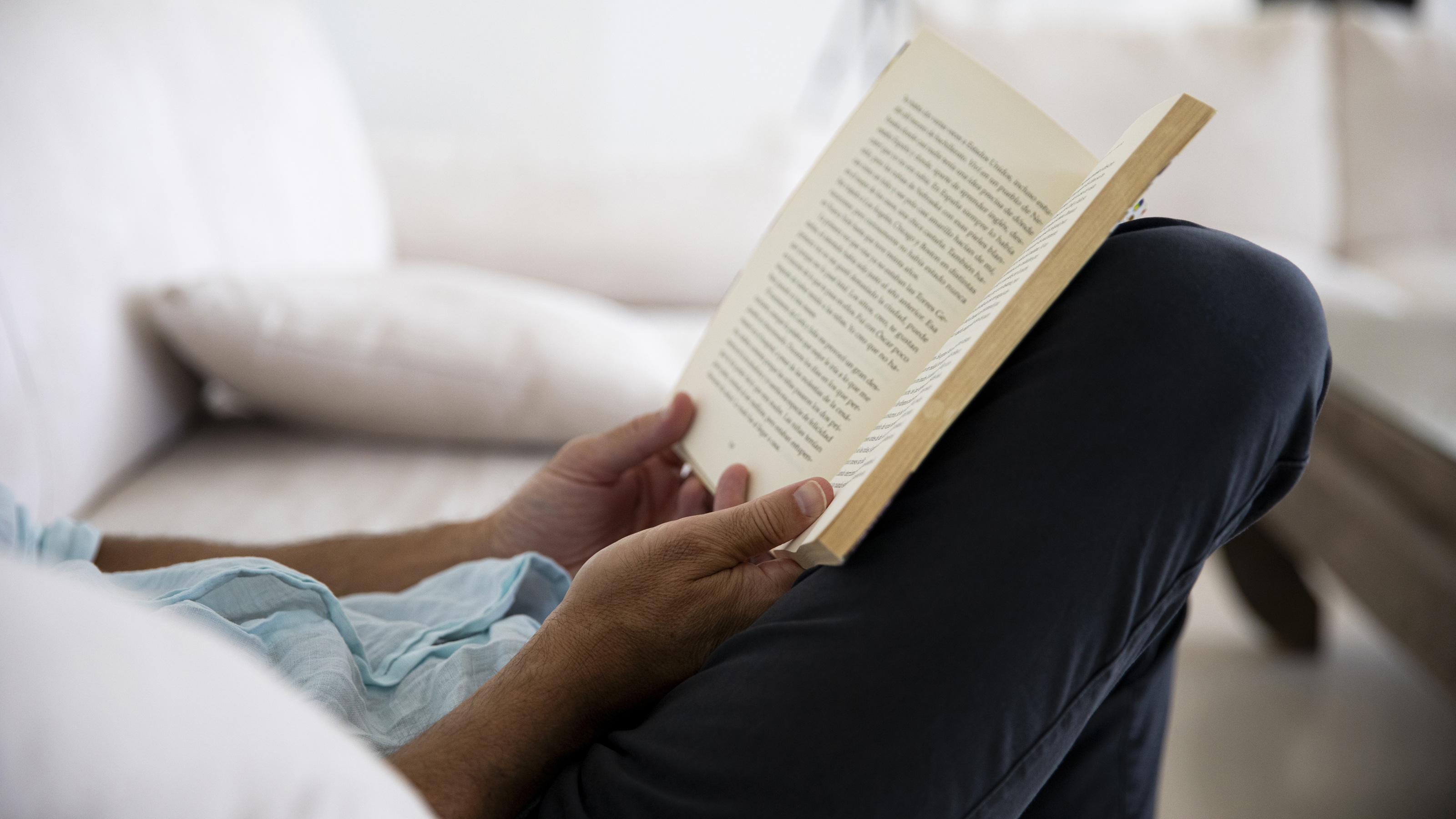 A man sits on the sofa with a book open on his lap.