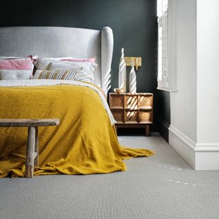 small bedroom colour ideas, grey upholstered bed with yellow throw, wooden stool at end, grey stripe carpet, charcoal black wall, white shutters, pops of pink pillows, wood side table, vases