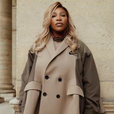 Serena Williams stands in front of a wall in Paris wearing a Nike camel trench coat