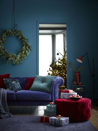 Christmas living room, blue walls, lilac couch, claret foot stool, side table, red floor lamp, purple rug, gifts, large wreath on the wall, Christmas tree in background, jade green and claret cushions