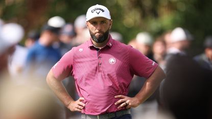 Jon Rahm with his hands on his hips while wearing a pink polo shirt and white cap
