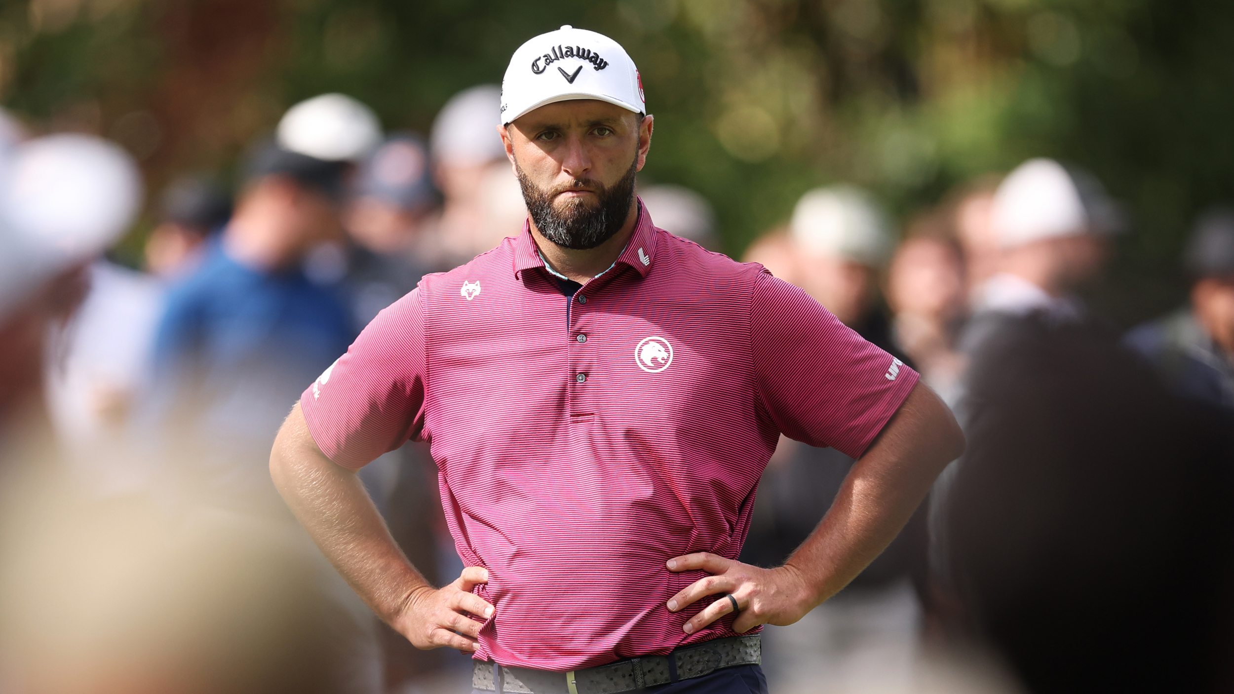 Jon Rahm with his hands on his hips while wearing a pink polo shirt and white cap