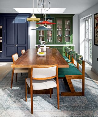 Dining room with mid-century, dark wooden dining table and chairs, upholstered in green, cream and patterned fabric, large blue and cream patterned rug, four different color and shaped pendant light hanging low above the table, painted blue and green storage and display cabinets